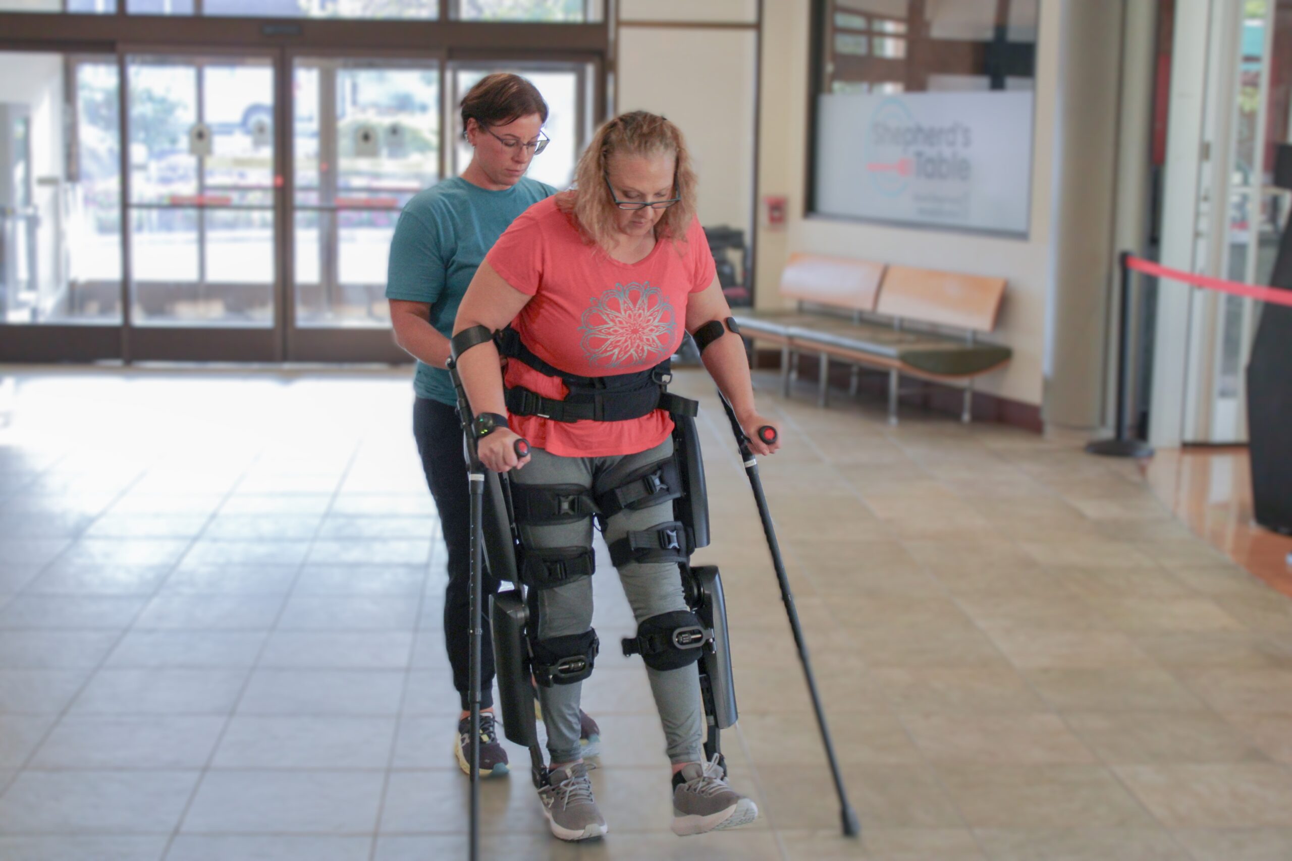 A person walks in a robotic exoskeleton with a Good Shepherd clinician standing behind them